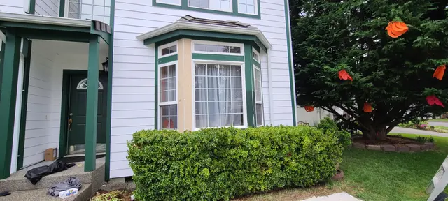 White house with green trim, covered porch and bay window beside a red maple